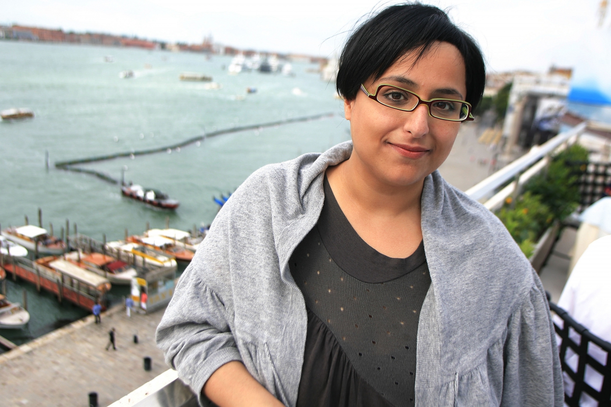 Woman wearing glasses, a black top, and a gray shawl standing near a waterfront with boats in the background.