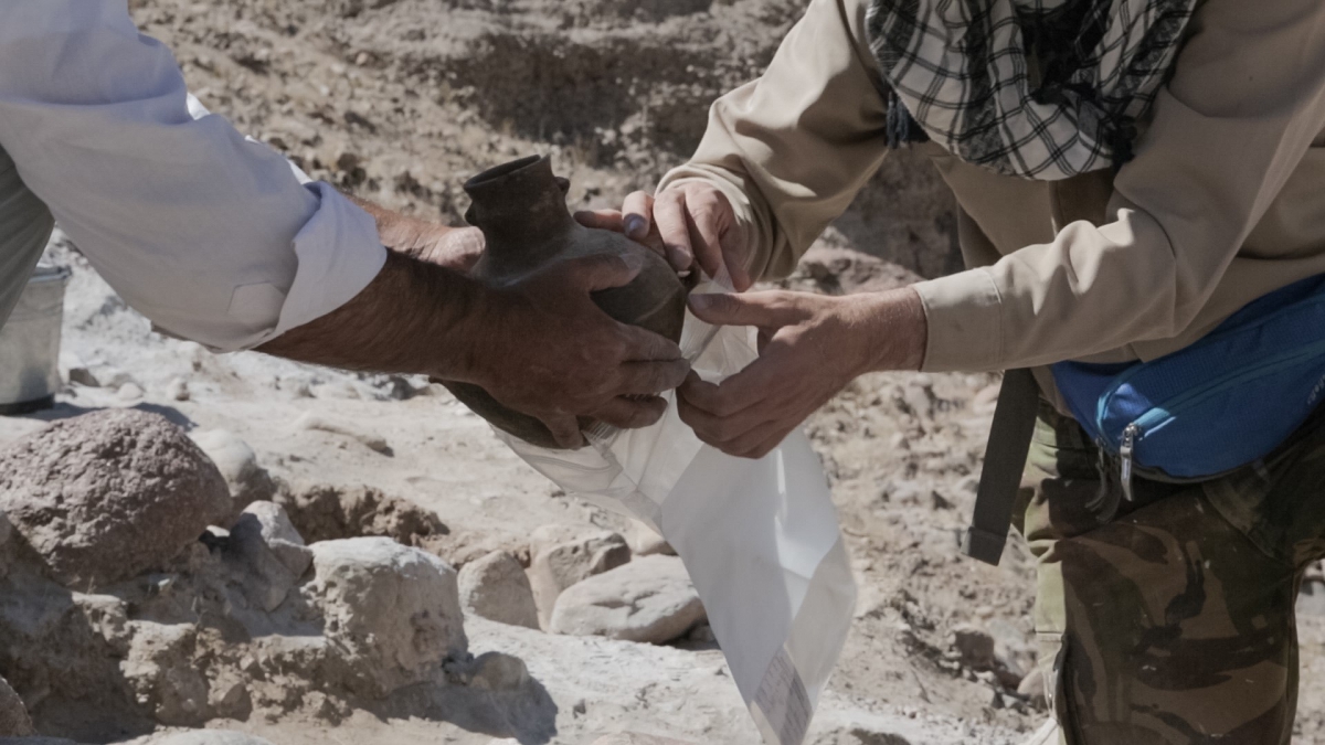 Two people carefully handling a dark brown pottery artifact over a sandy excavation site.