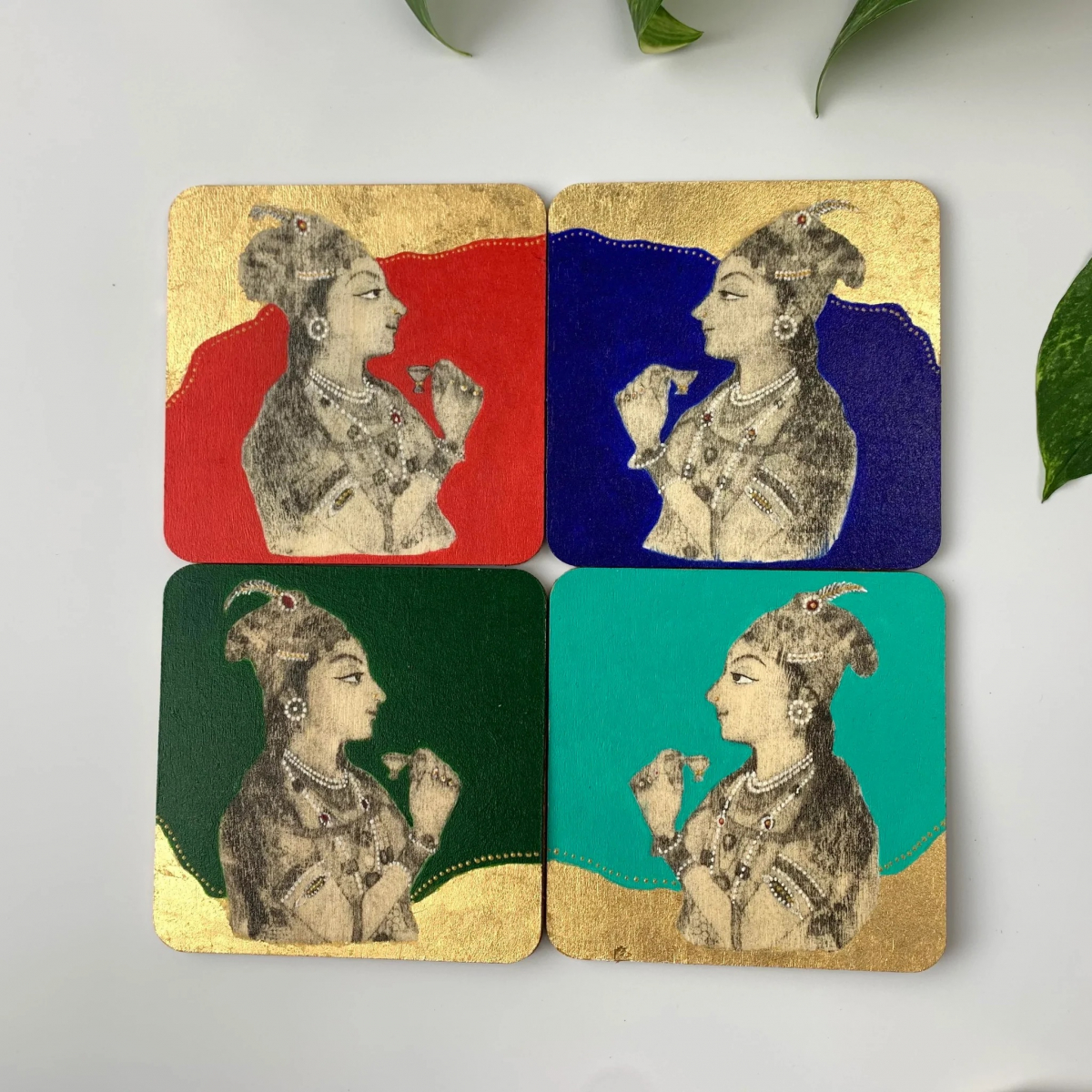 Four square coasters each featuring a stylized profile of a woman wearing a headpiece and holding a flower, with red, blue, dark green, and teal backgrounds.