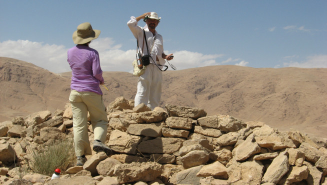 Two people wearing hats stand among rocks in a dry, mountainous landscape under a blue sky.