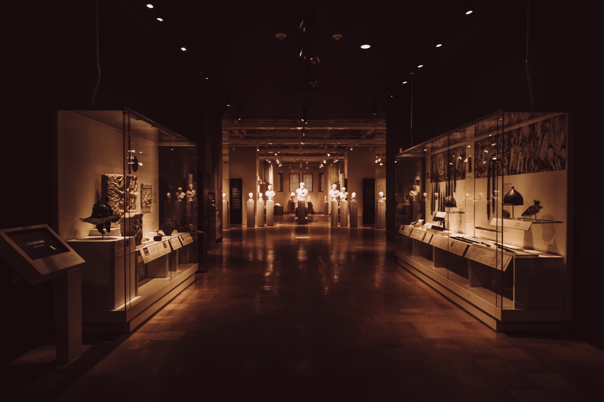 Museum gallery interior with dim lighting, featuring glass display cases holding various artifacts along both sides.