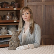 A woman wearing white gloves poses with an ancient carved stone artifact on a table, with wooden shelves holding pottery in the background.