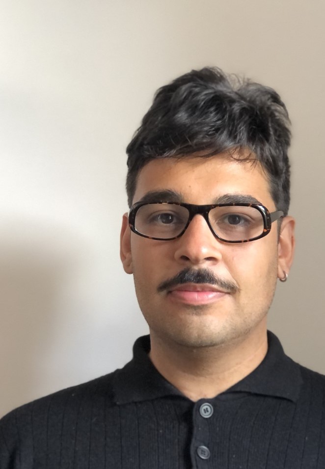 Portrait d'un homme aux cheveux bruns et courts, portant des lunettes, une fine moustache et une chemise noire sur un fond clair et uni.