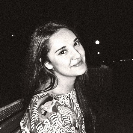 Young woman with long dark hair smiling and wearing a patterned top, photographed in black and white at night.