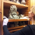 A woman wearing white gloves carefully examines a detailed carved stone artifact on a wooden shelf.