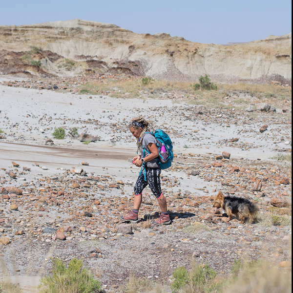 A man with a blue backpack walks with a small dog on a rocky desert landscape with eroded cliffs.