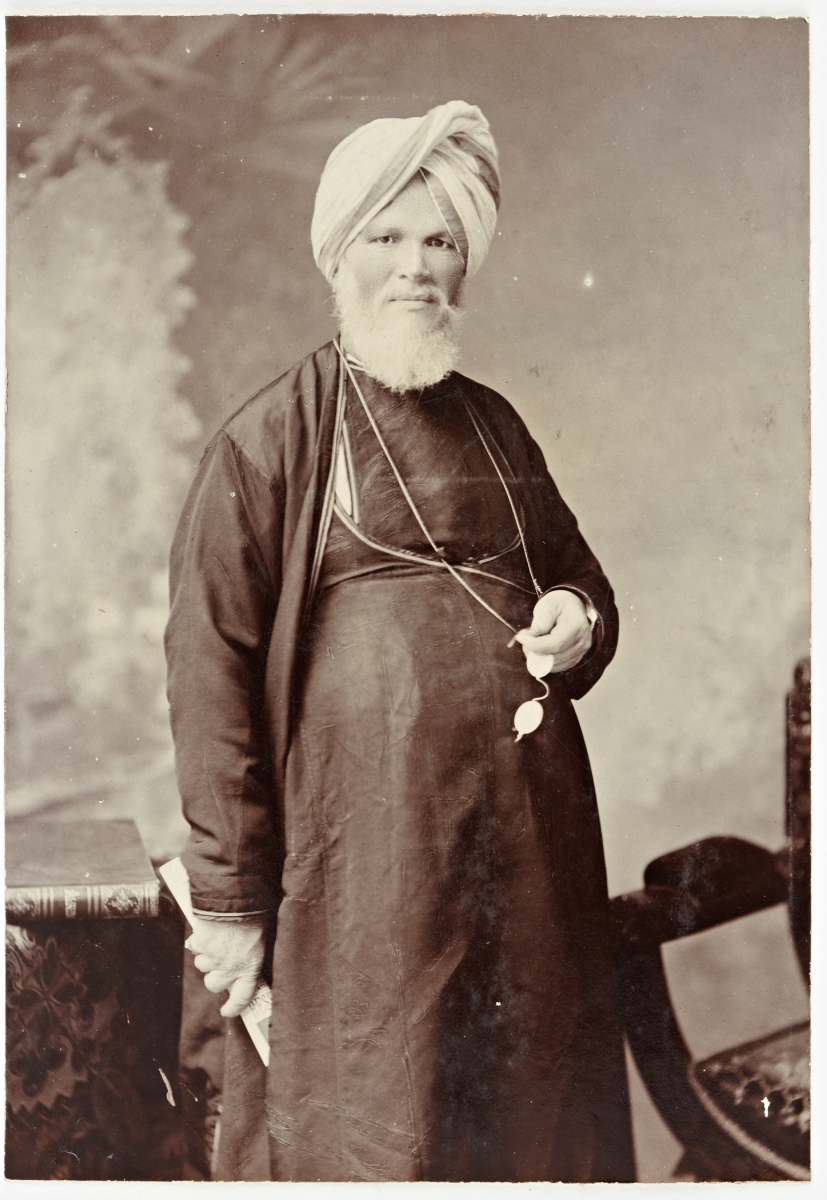 Elderly man wearing a turban and traditional attire, standing near a chair in a sepia-toned portrait.