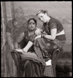 Two women in traditional Indian attire, one seated and the other leaning on her knee, posed against a plain backdrop.