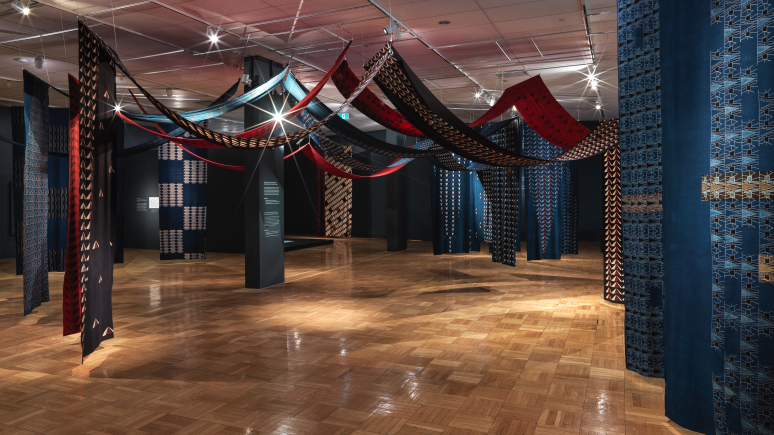 Installation composée de panneaux de tissu rouge, noir et bleu suspendus au plafond d'une galerie spacieuse dotée de parquets en bois.
