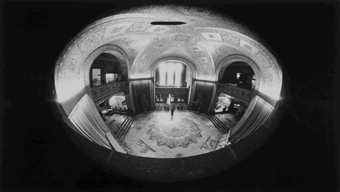 No visible text; black-and-white fisheye lens photo showing a circular room with large arched windows and a patterned floor.