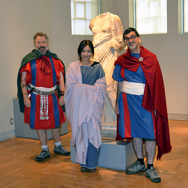 Three people wearing ancient Roman-style clothing stand inside a museum gallery with a Roman statue in the background.