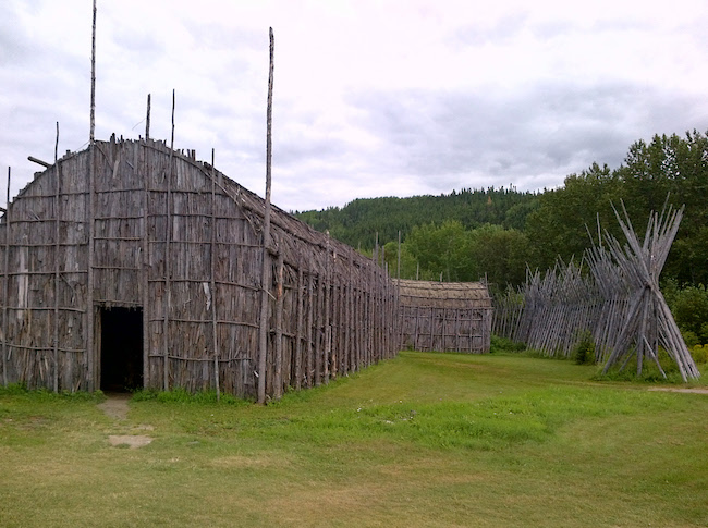 Large wooden longhouse structure with a curved roof made of vertical wooden slats, standing on grassy ground with trees and hills in the background.