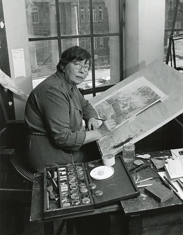 A woman seated at a desk painting on a large angled drawing board with paint and brushes nearby.