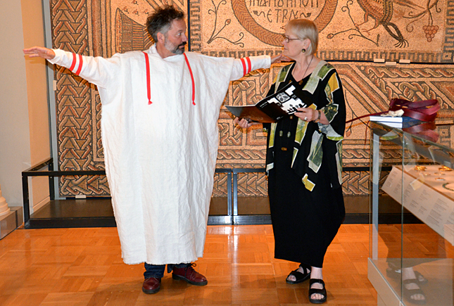 Two people standing in a museum gallery, one wearing a large white garment with red ties, and the other in a black outfit with patterned accents, smiling and interacting.