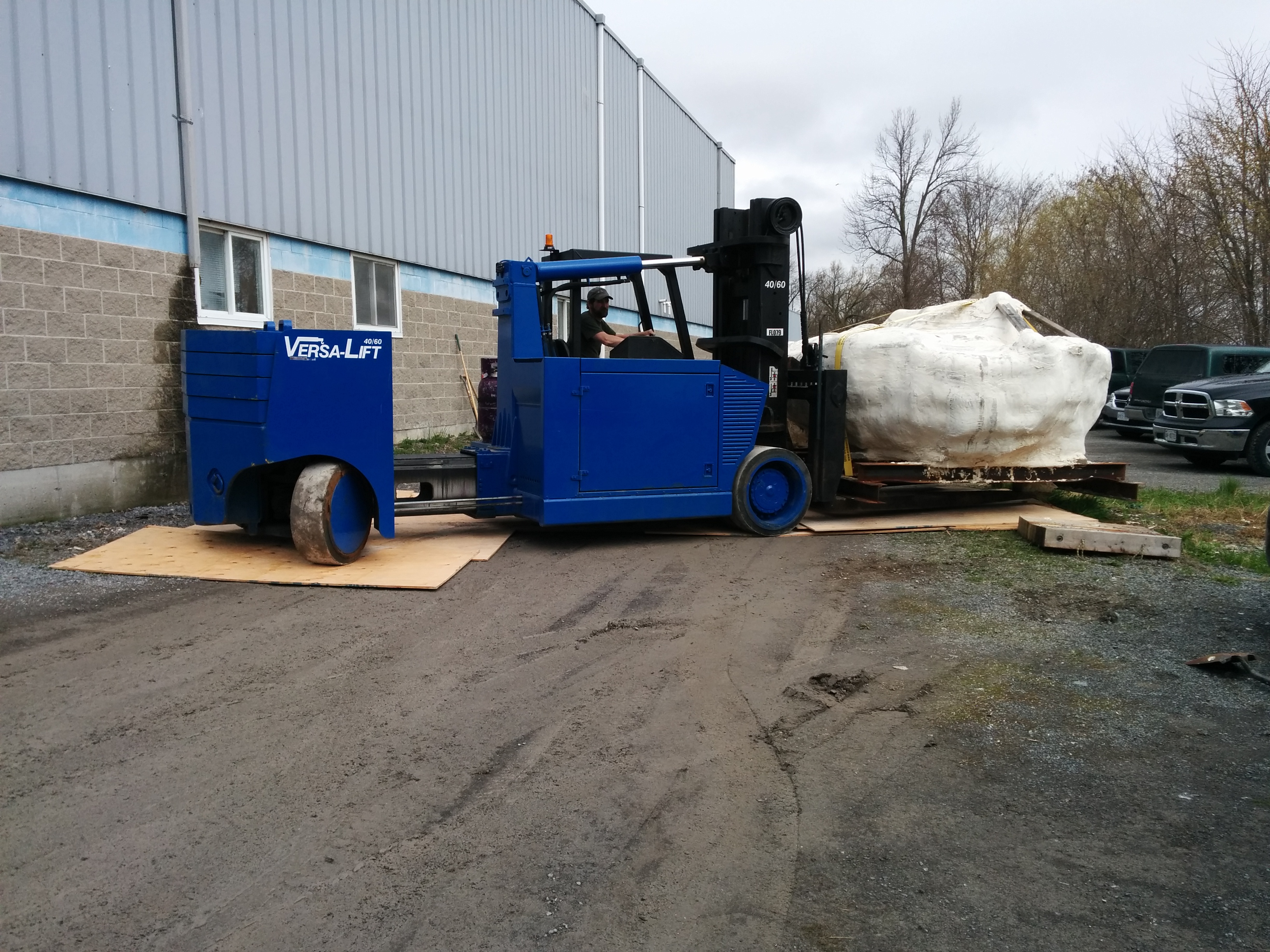 Blue forklift carrying a large, white wrapped object outside a gray industrial building.