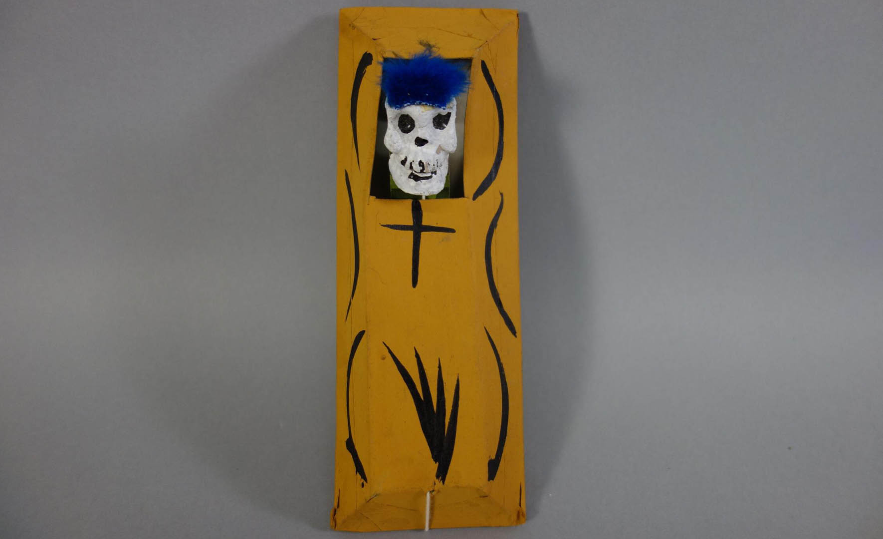 Objet rectangulaire jaune avec des lignes courbes noires et un petit crâne blanc au sommet.