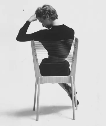 Person sitting on a wooden chair facing backward with their left arm raised to their forehead against a plain white background.