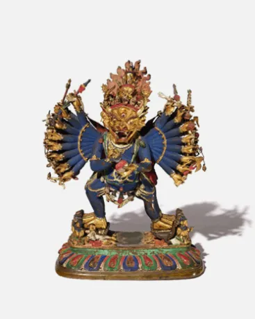Statue of a multi-armed deity with a fierce expression, adorned with gold and blue garments, standing on a decorated platform.
