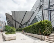 Modern angular glass and metal building facade with geometric design and greenery along a city sidewalk.
