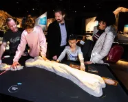 Five people, including a child, examine a large dinosaur bone displayed on a black table in a dimly lit museum gallery.
