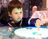 A young boy with dark hair interacts with a circular touchscreen display, with exhibit items blurred in the background.