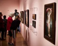 Several people closely observing framed artworks displayed on a museum wall with soft pink lighting.