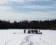 Groupe de personnes rassemblées sur un lac gelé et enneigé, entouré d'arbres, sous un ciel partiellement nuageux.