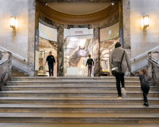 Grand marble staircase inside a building with people ascending and descending, flanked by ornate walls and sconces.