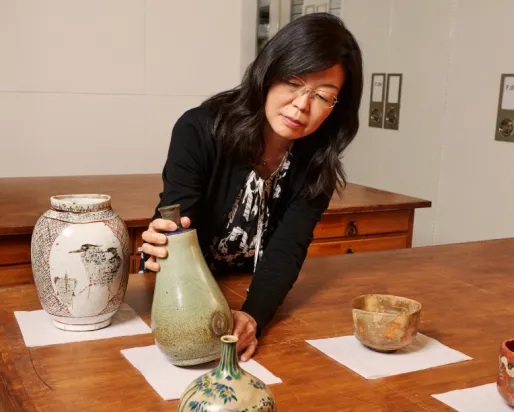 Woman arranging four ceramic vessels of different shapes and sizes on a wooden table against a plain wall.
