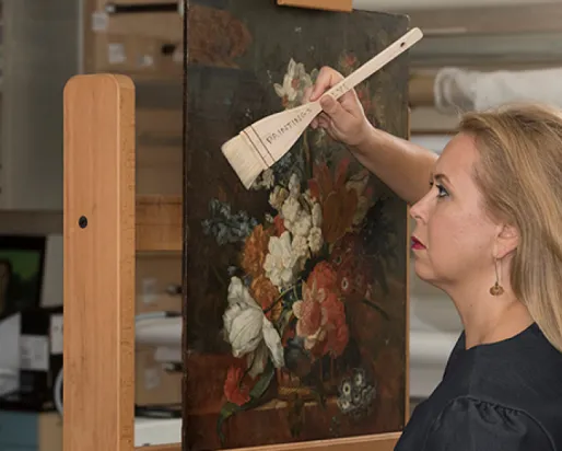 Woman holding a wooden hand fan in front of a painting of a floral bouquet.