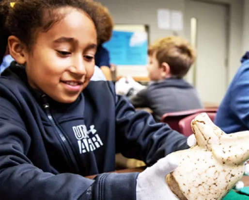 Girl in a dark shirt holding a small speckled artifact shaped like an animal, with a blurred classroom background.