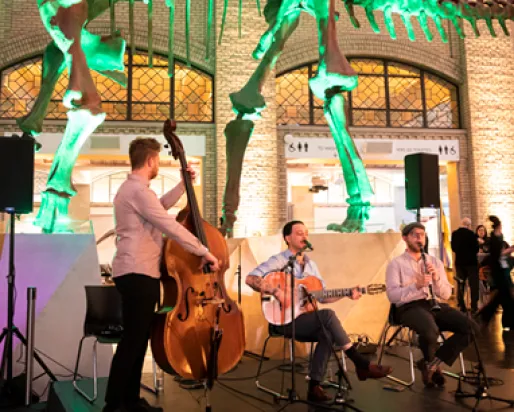 A three-member band performs inside a museum under a large dinosaur skeleton with green lighting.
