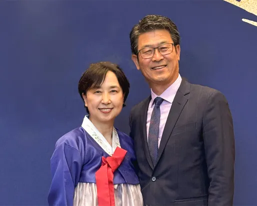 A man in a dark suit and glasses stands next to a woman wearing a traditional Korean hanbok dress with a blue top and red bow.