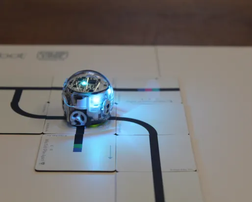 A small round robot with blinking lights follows a dark path along a paper