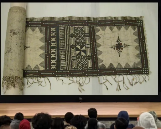 Une femme indigène souriante se tient derrière un podium sur une scène. Sur un écran à côté d'elle, on voit l'image d'un morceau de tissu avec des motifs complexes à l'encre noire et brune.