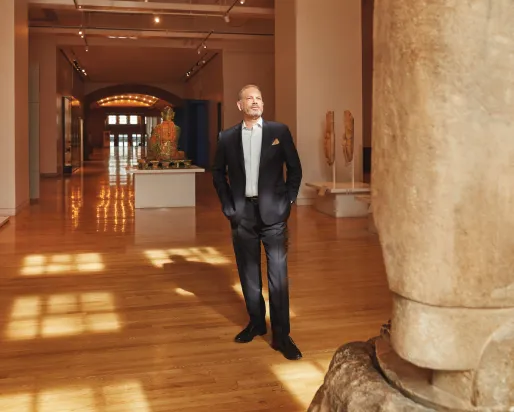 A man in a black tuxedo standing in a spacious gallery with wooden floors, stone columns, and display cases.