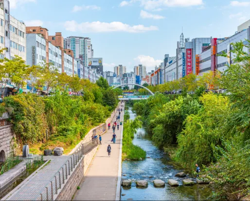 Urban park with a walking path, railway tracks on the left, trees, and a river flowing through the middle under a blue sky.