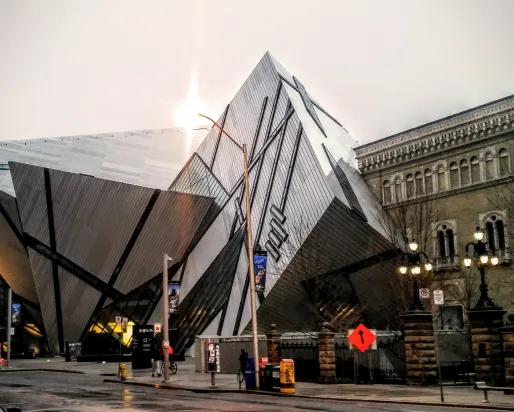 Modern angular glass and metal building facade of the Royal Ontario Museum with sunlight behind it, and a historic building in the background.