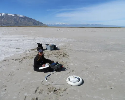 Researcher sitting on a dry lakebed next to scientific equipment including a circular white sensor device.