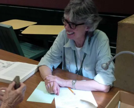 A woman sitting at a desk writes on paper with open books and a black pen visible on the table.