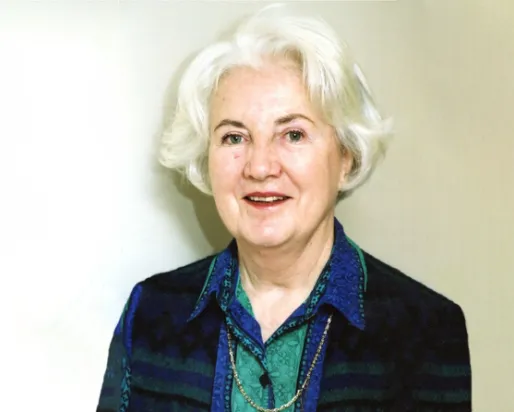 Portrait of an older woman with white hair, wearing a dark vest and a blue patterned blouse against a white background.