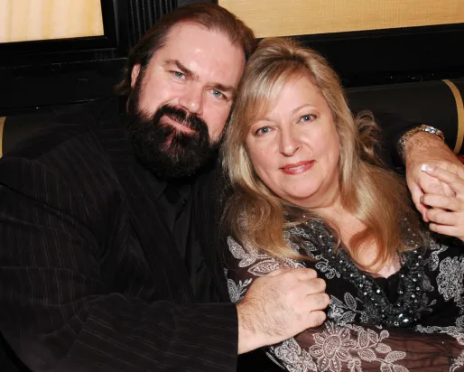A man with a beard and a woman with blonde hair sit together on a striped couch, both looking at the camera.