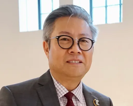 Man with glasses wearing a dark suit and red patterned tie, standing indoors against a beige wall.