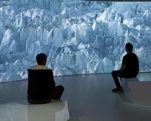 Two people sit on benches facing a large digital wall displaying a blue-hued glacier scene.