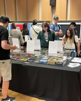 Several people viewing a table covered in a selection of gaming consoles
