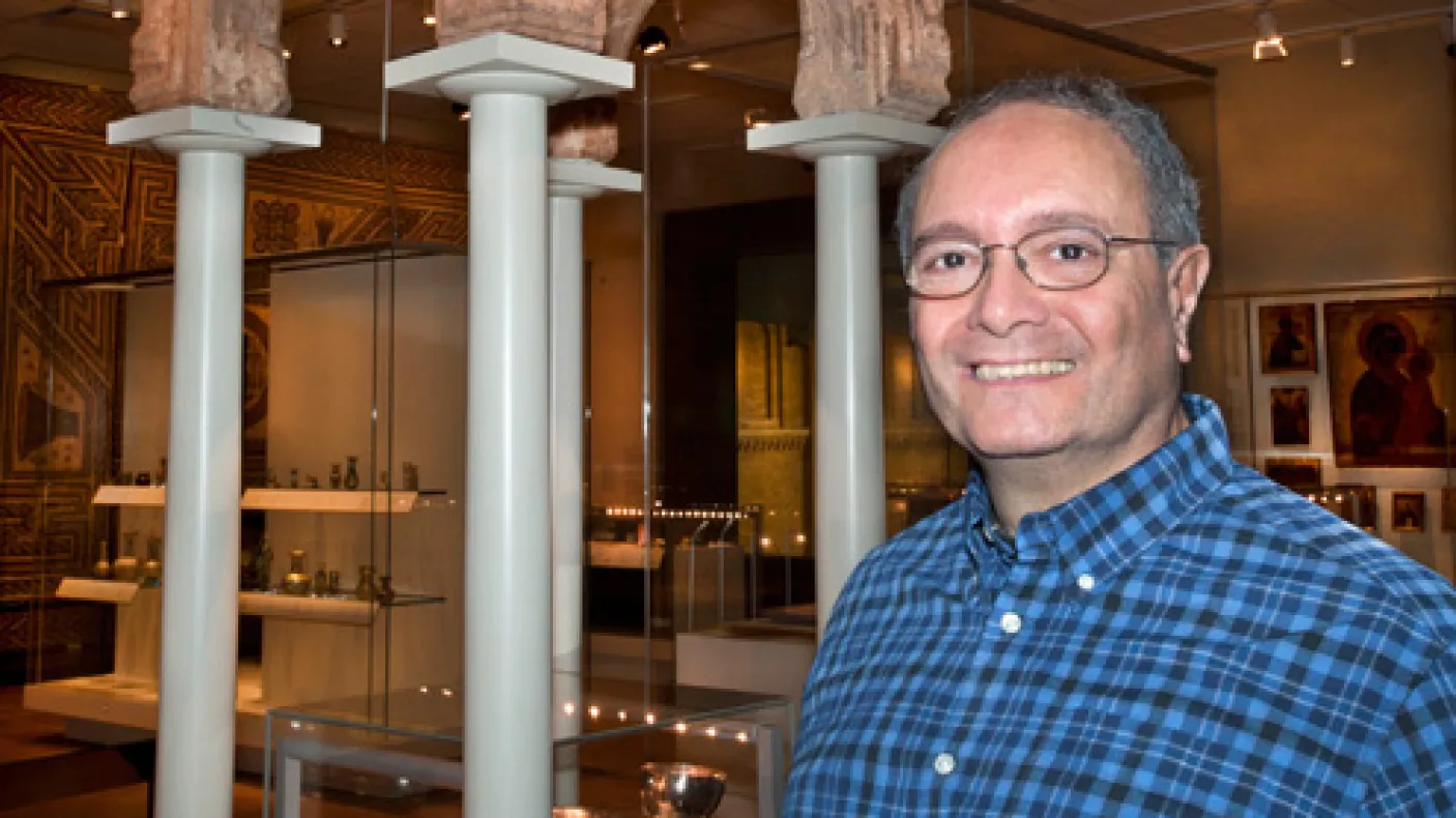 A man wearing glasses and a blue checked shirt stands in a museum gallery with white columns and display cases behind him.
