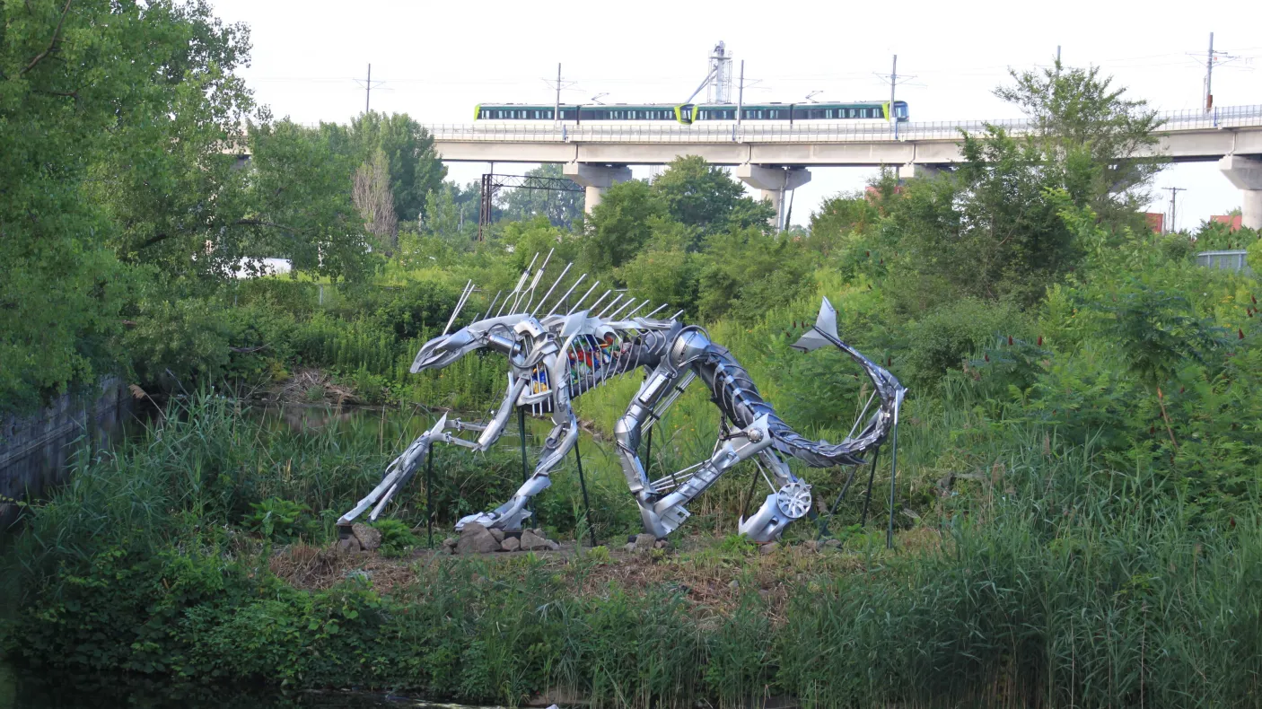 Sculpture en acier représentant un chat à dents de sabre accroupi, entouré de verdure, près d'un plan d'eau, avec un train qui passe à l'arrière-plan.