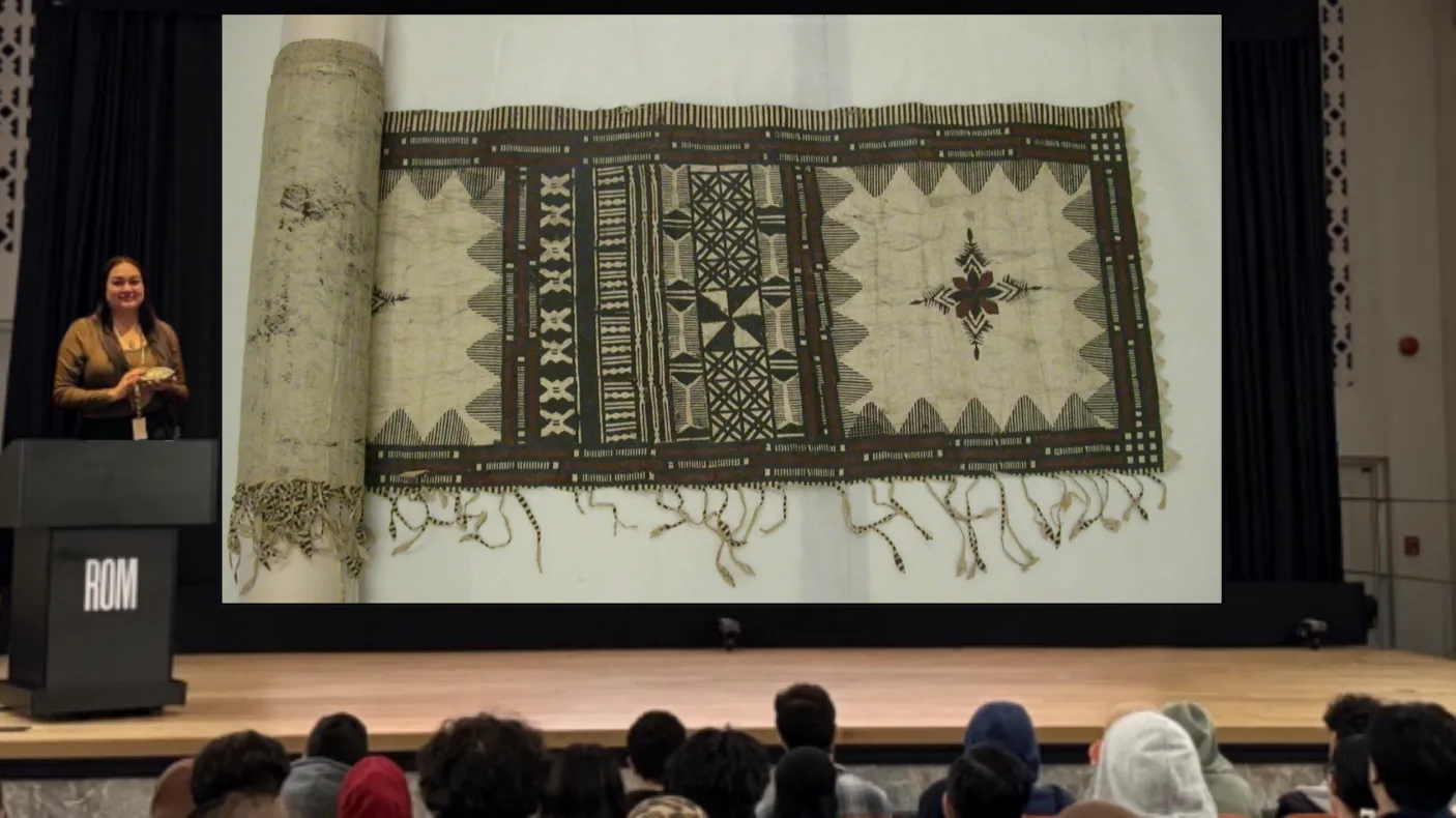 A smiling Indigenous woman stands behind a podium on a stage. On a screen next to her is an image of a length of cloth with intricate patterns in dark black and brown ink.