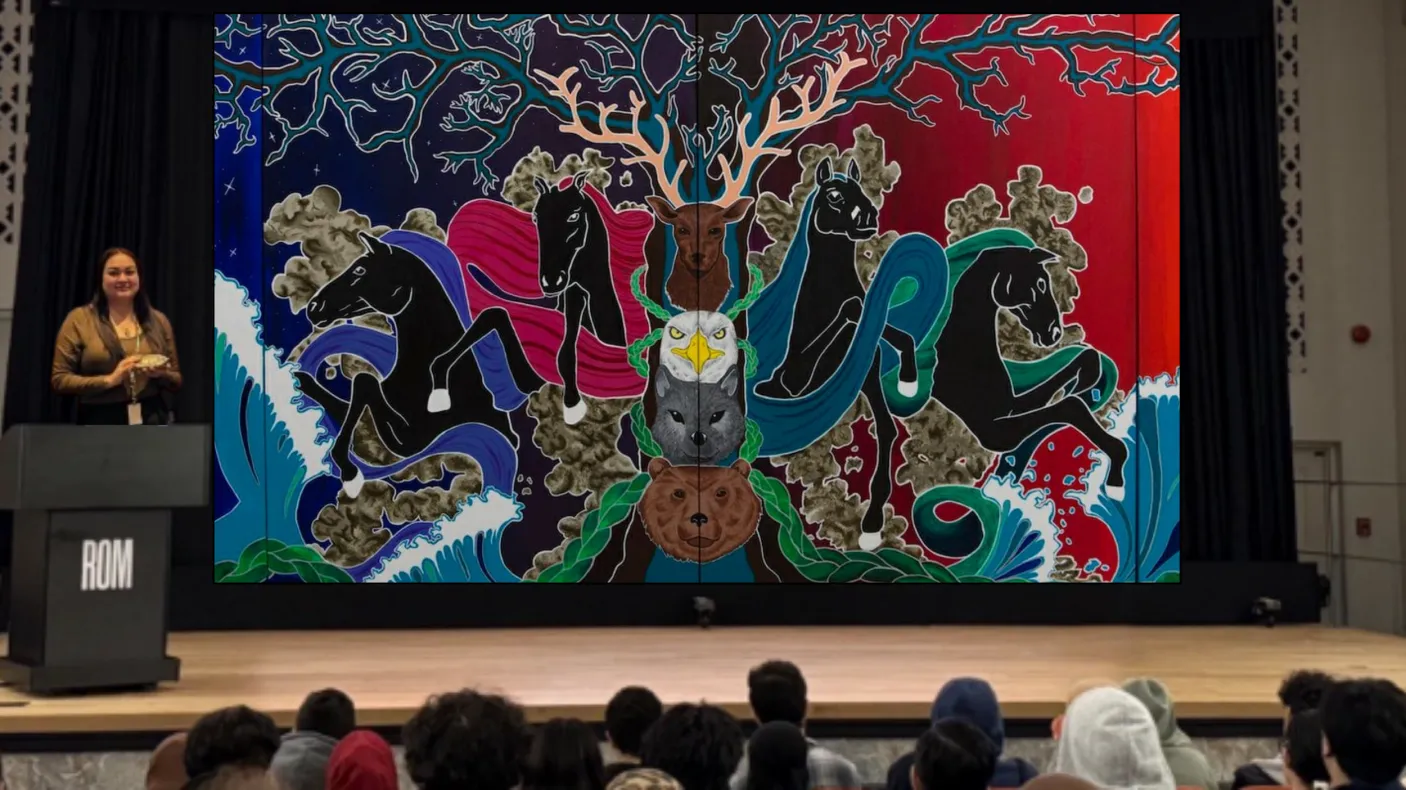 An Indigenous woman stands on a stage behind a podium. A screen behind her displays a painted mural.