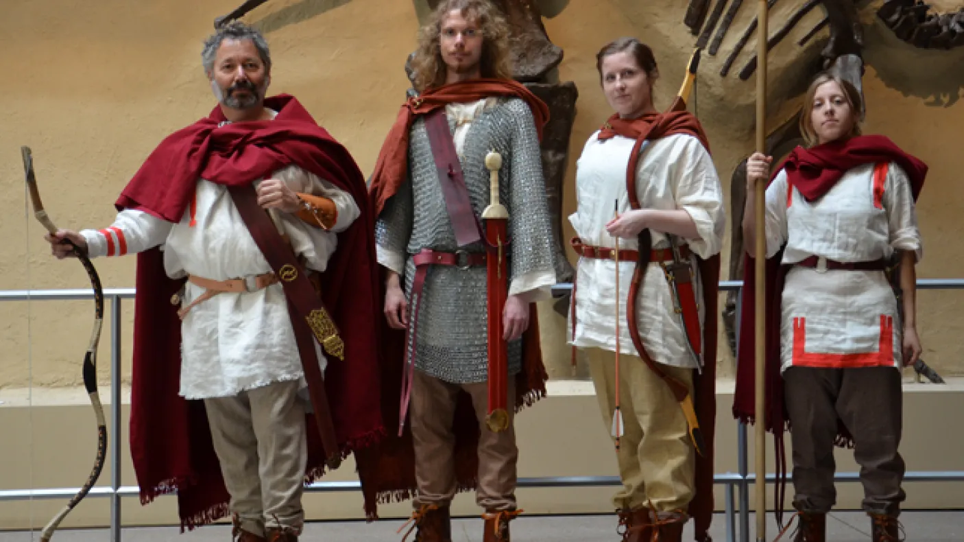 Four people dressed in ancient Roman soldier costumes standing indoors with a large dinosaur fossil mounted on a wall behind them.